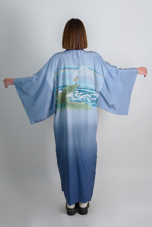 May include: A blue silk kimono with a mountain and ocean scene printed on the back. The kimono has a gradient color from light blue to dark blue.