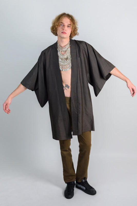 May include: A person wearing a dark brown kimono with a silver layered necklace and brown pants. The person is standing on a white background.