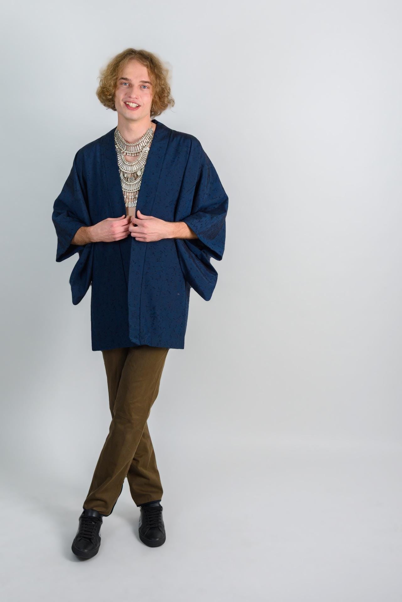 May include: A person wearing a dark blue kimono with a silver necklace and brown pants. The person is standing in front of a white background.