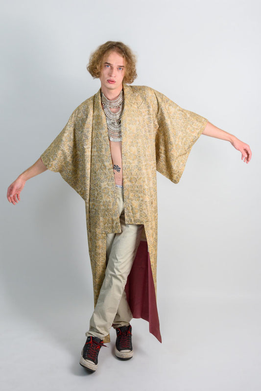 May include: A person wearing a long, gold and burgundy patterned robe with a silver necklace. The robe is open in the front, revealing a pair of beige pants. The person is wearing black and red sneakers.