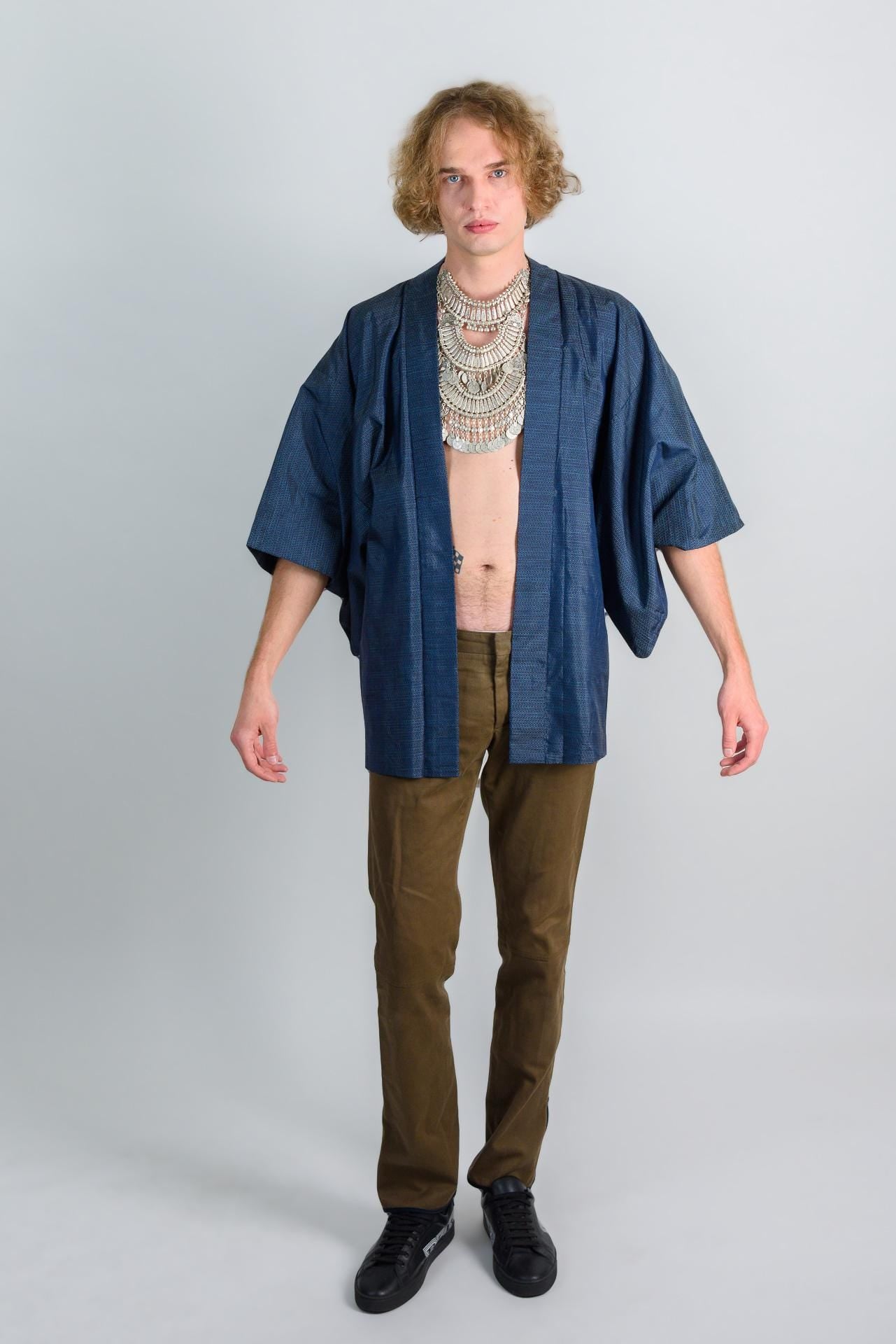 May include: A person wearing a blue kimono with a silver necklace and brown pants.