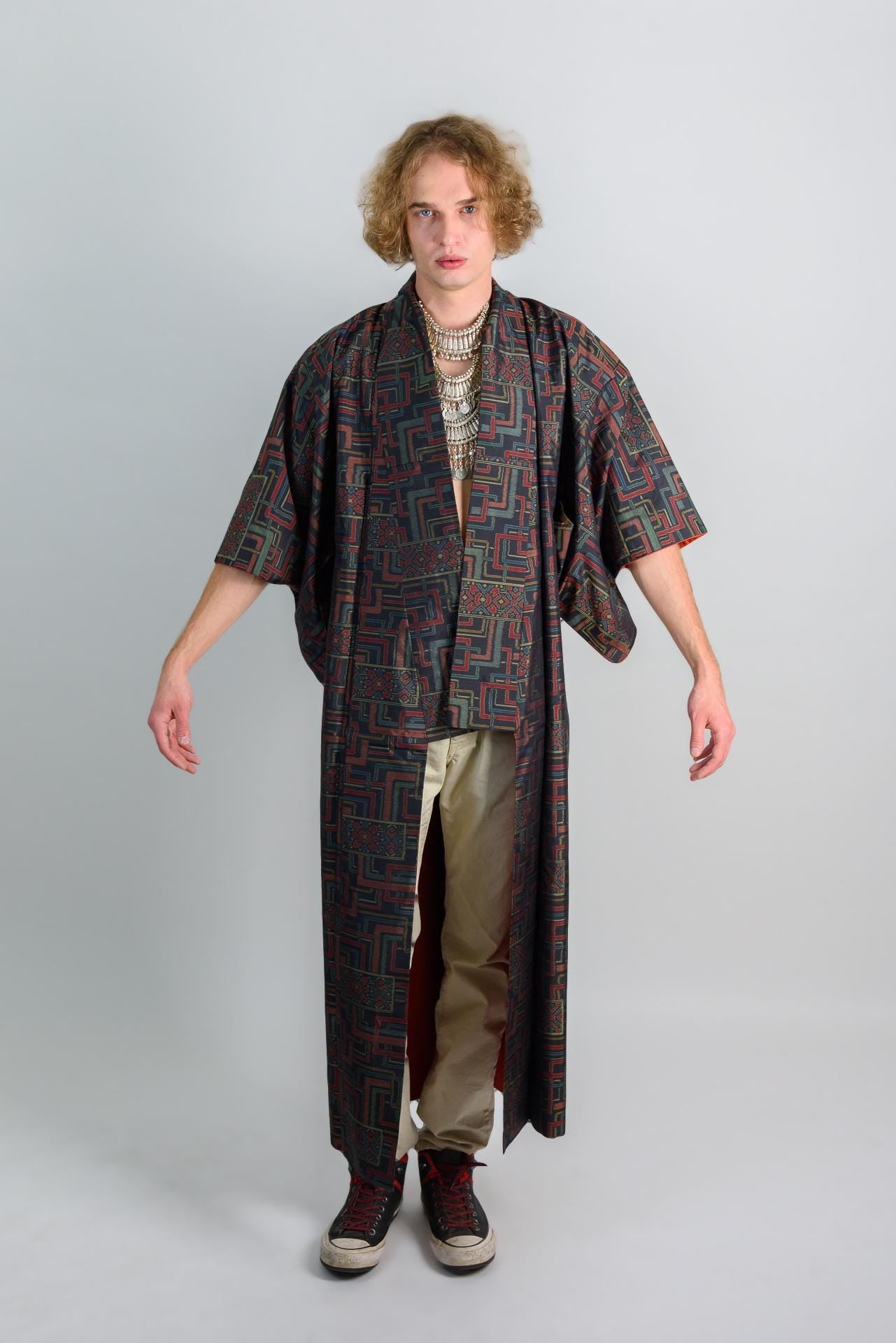 May include: A person wearing a long, black and red patterned robe with a silver necklace. The robe has long sleeves and a wide collar. The person is wearing beige pants and black shoes.