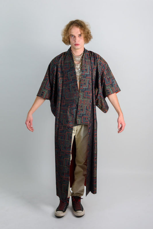 May include: A person wearing a long, black and red patterned robe with a silver necklace. The robe has long sleeves and a wide collar. The person is wearing beige pants and black shoes.