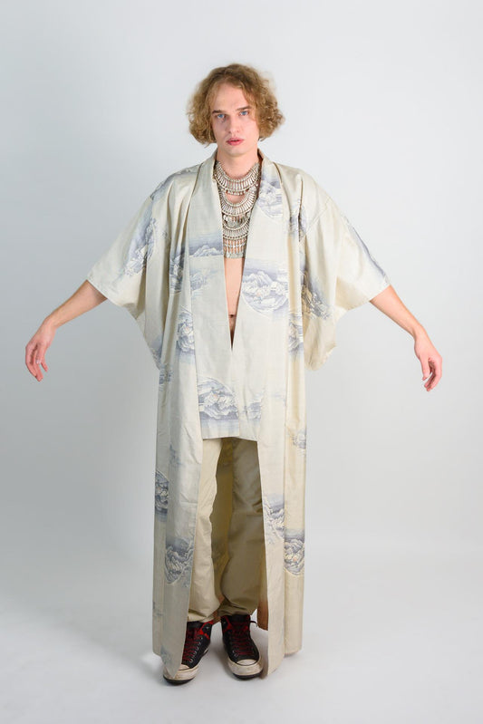 May include: A white kimono with a blue and white mountain pattern. The kimono has long sleeves and a wide, open collar. The kimono is worn over a pair of beige pants and black shoes.