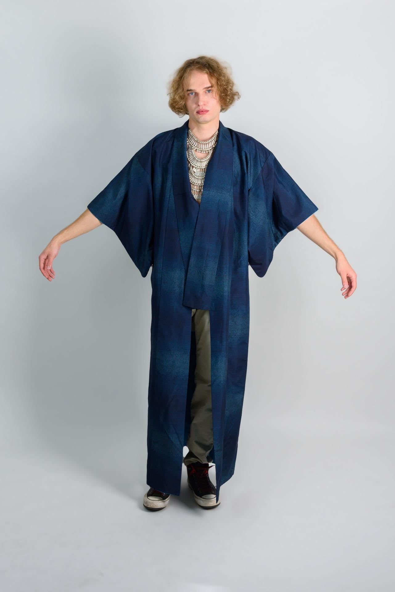 May include: A person wearing a long, dark blue kimono with a silver necklace. The kimono has a subtle pattern of light blue and dark blue stripes. The person is wearing black sneakers.