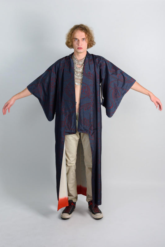 May include: A blue and red patterned kimono with long sleeves. The kimono has a white and red trim at the bottom. The person is wearing beige pants and black shoes.