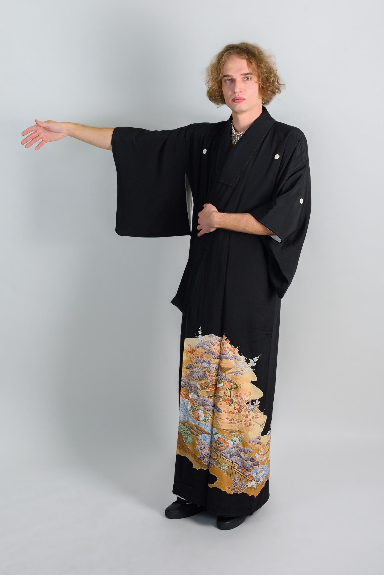 May include: A person wearing a black kimono with a colorful floral pattern on the bottom half. The kimono has long sleeves and is tied at the waist. The person is standing with their left arm extended and their right hand on their hip.