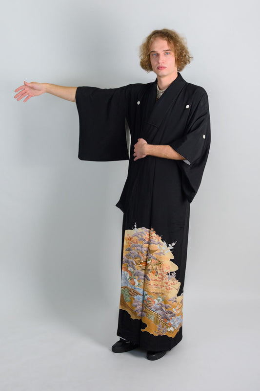 May include: A person wearing a black kimono with a colorful floral pattern on the bottom half. The kimono has long sleeves and is tied at the waist. The person is standing with their left arm extended and their right hand on their hip.