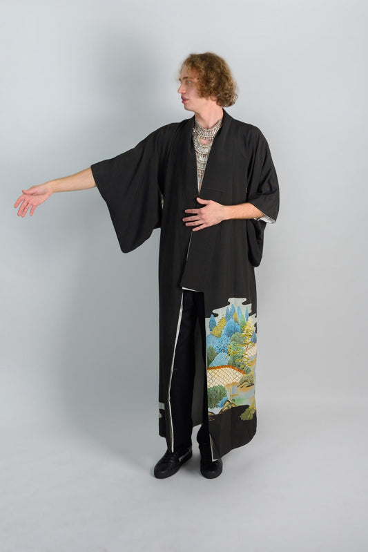 May include: A person wearing a black kimono with a colorful landscape print on the bottom right side. The kimono has a long sleeve and is open in the front. The person is wearing black pants and black shoes.