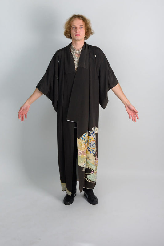 May include: A person wearing a black kimono with a floral pattern on the bottom. The kimono has a long sleeve and is open in the front. The person is wearing black shoes.