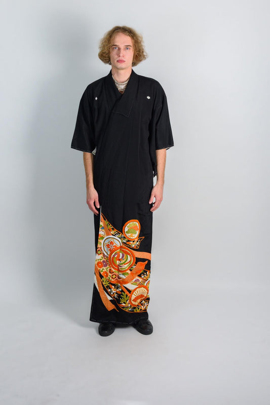 May include: A person wearing a black kimono with orange and white floral patterns. The kimono has a long sleeve and a wrap-around style. The person is standing on a white background.