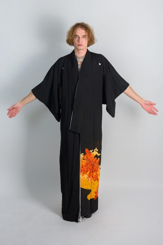 May include: A black kimono with a floral pattern on the bottom. The pattern is orange and yellow. The kimono is worn by a person with long blonde hair.