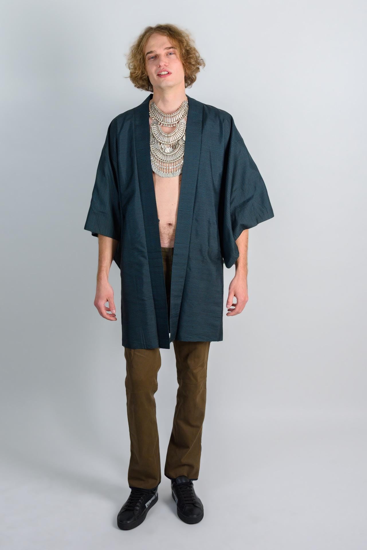 May include: A dark green kimono with a long sleeve and a simple design. The kimono is worn over brown pants and black shoes.