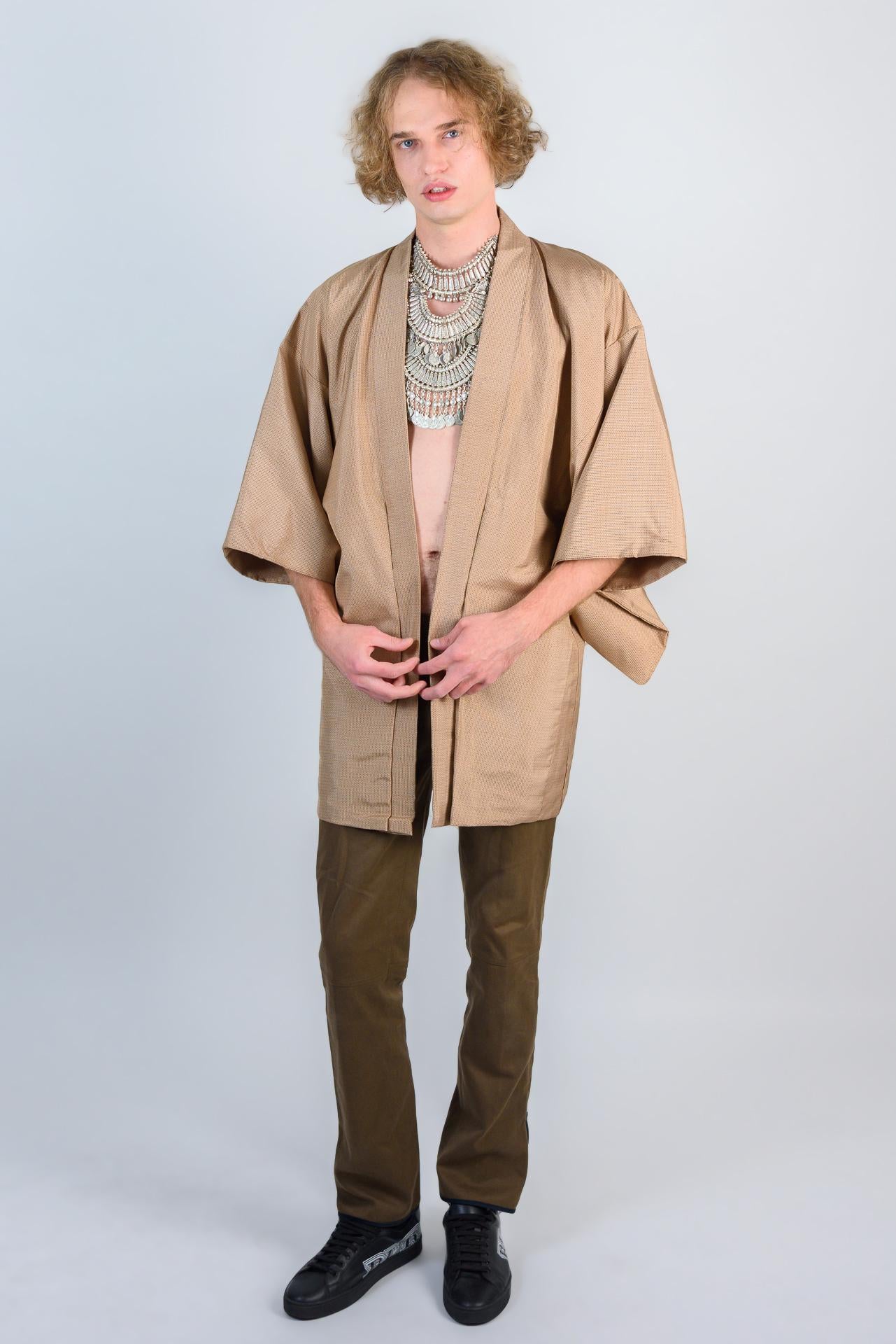 May include: A person wearing a tan kimono with a silver necklace and brown pants