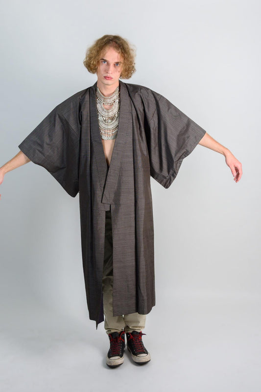 May include: A long, dark gray kimono with a black and white patterned design. The kimono is open in the front and has long sleeves. The person wearing the kimono is wearing a silver necklace with multiple layers.
