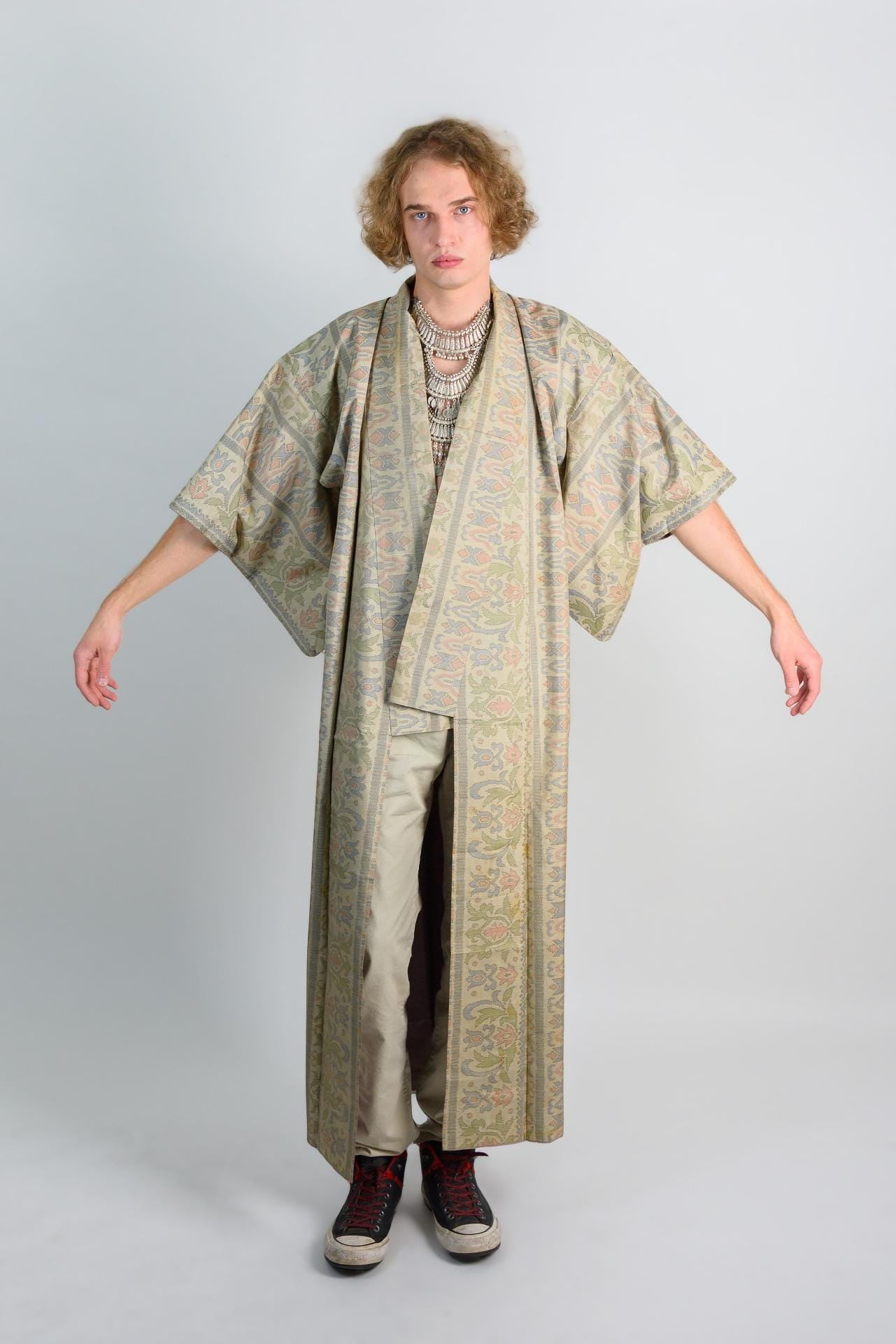 May include: A long, patterned robe with a floral design in shades of green, brown, and pink. The robe has a wide collar and long sleeves. The person is wearing a silver necklace and black shoes.