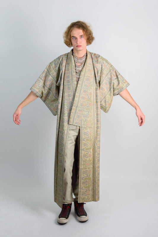 May include: A long, patterned robe with a floral design in shades of green, brown, and pink. The robe has a wide collar and long sleeves. The person is wearing a silver necklace and black shoes.