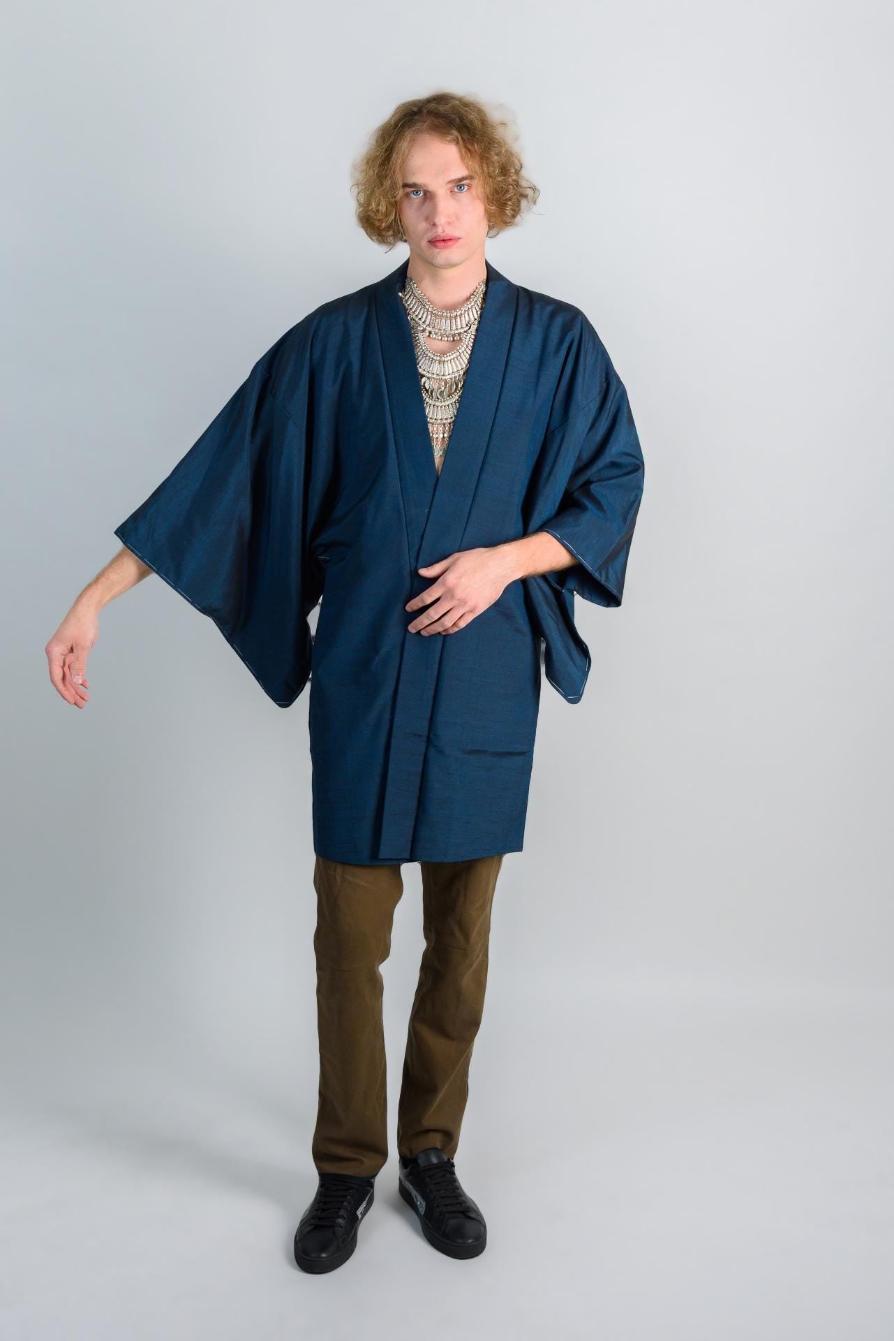 May include: A person wearing a dark blue kimono with a silver necklace and brown pants. The person is standing on a white background.