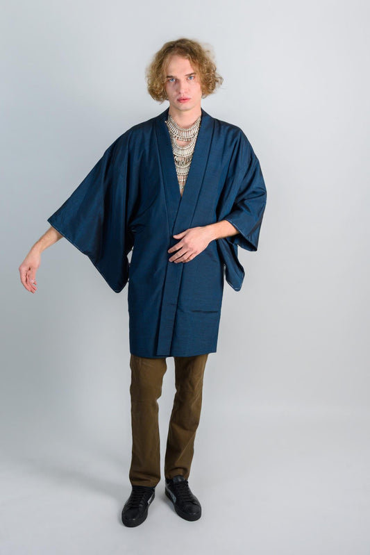 May include: A person wearing a dark blue kimono with a silver necklace and brown pants. The person is standing on a white background.