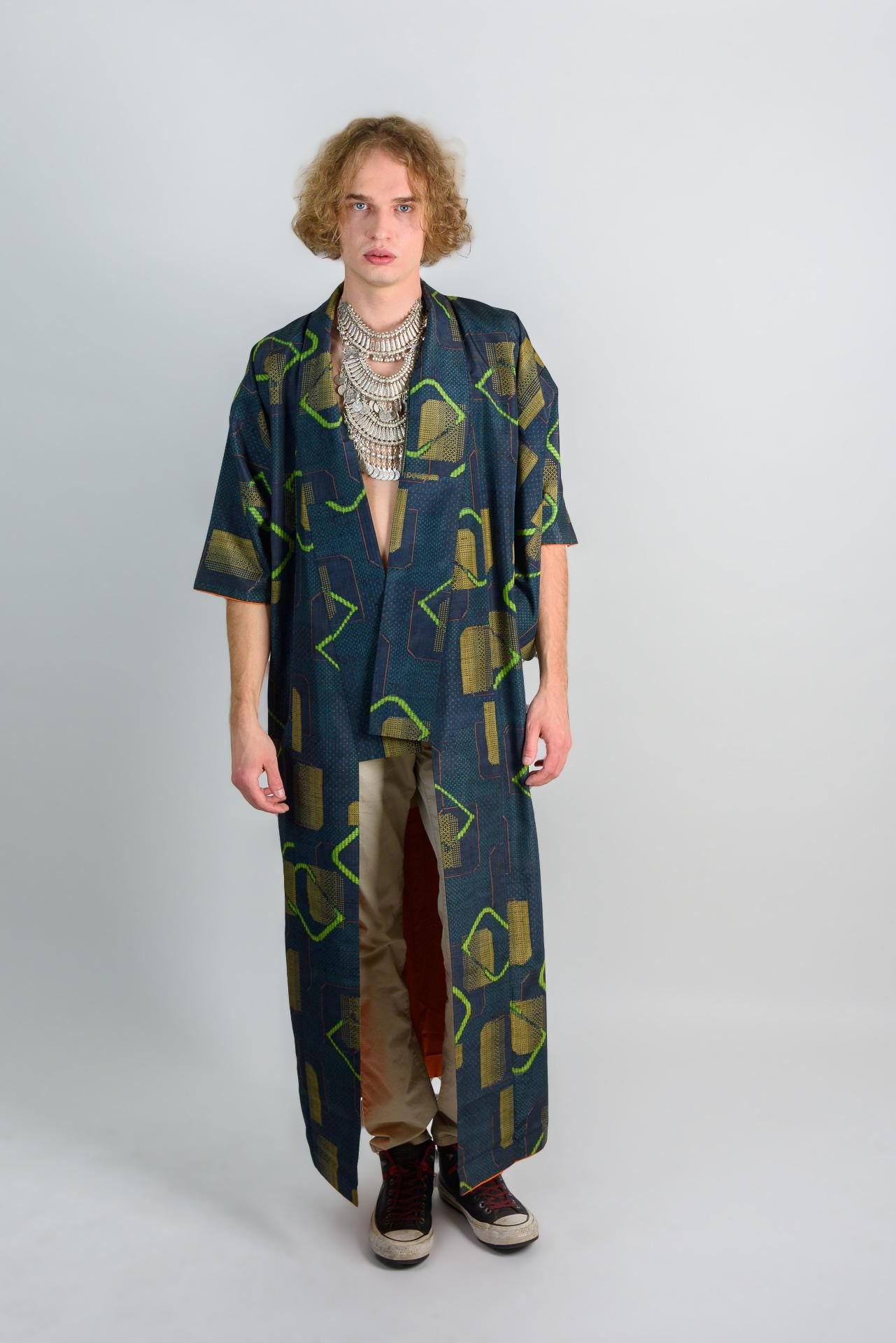 May include: A long, dark blue robe with a geometric pattern in green and yellow. The robe has a long, flowing silhouette and is open in the front. The person is wearing a silver necklace and has long, curly hair.