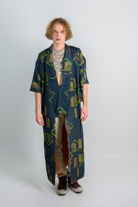 May include: A long, dark blue robe with a geometric pattern in green and yellow. The robe has a long, flowing silhouette and is open in the front. The person is wearing a silver necklace and has long, curly hair.