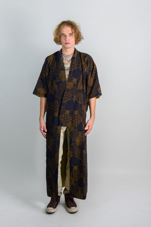 May include: A person wearing a long, dark blue and gold patterned robe with a silver necklace. The robe has a geometric pattern and is open in the front. The person is wearing beige pants and black shoes.