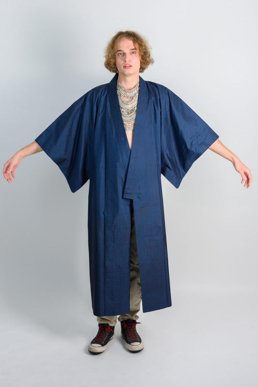 May include: A long, dark blue kimono with a long, open front and long sleeves. The kimono is made of a soft, textured fabric. The kimono is worn over a pair of light brown pants and black sneakers.