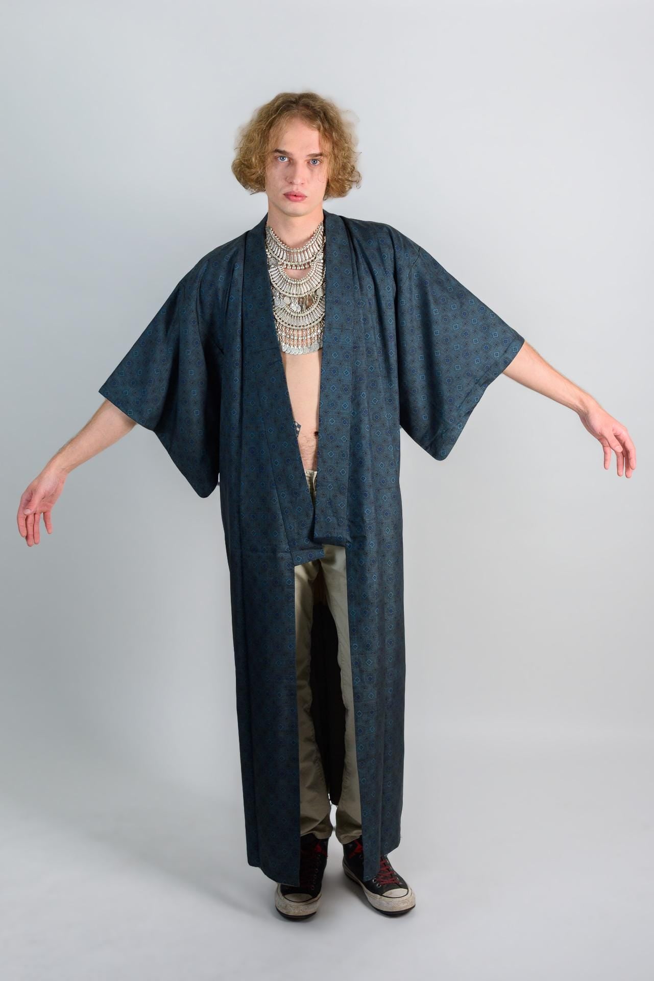 May include: A long, dark blue kimono with a small floral pattern. The kimono has long sleeves and a wide collar. The kimono is open in the front, revealing a white shirt underneath. The person is wearing a silver necklace with multiple layers.